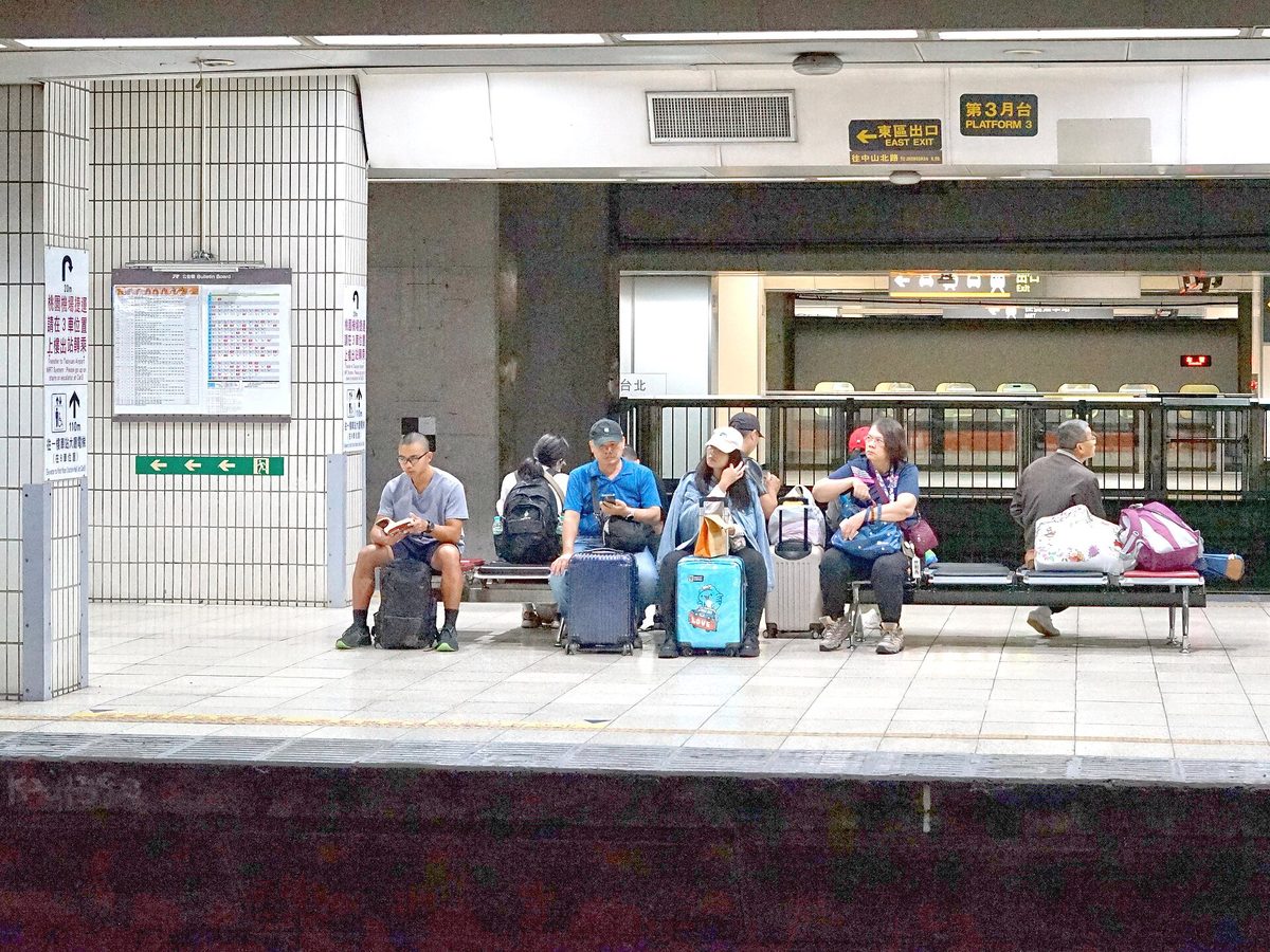 車站候車區長椅上坐著數名旅客，腳邊放著行李箱